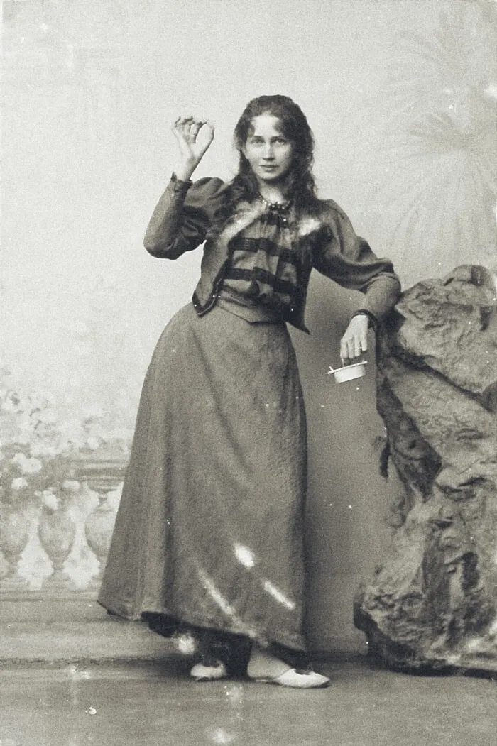 Black and white studio portrait of composer Tekla Griebel Wandall, circa 1896. She stands in a full-length dress, one hand raised with thumb and index finger pressed together, the other resting on a decorative prop. She looks slightly past the camera.